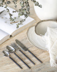 A minimalist table setting featuring hand-forged stainless steel cutlery with sleek black handles, paired with rustic ceramic dishware on a textured stone table, evoking a refined yet organic dining experience