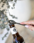 A hand-forged black iron bottle opener in use, effortlessly opening a Burleigh Bighead beer bottle against a stylish neutral-toned setting with a white ceramic vase and dried foliage