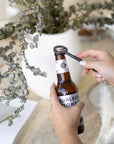 A hand-forged black iron bottle opener in use, effortlessly opening a Burleigh Bighead beer bottle against a stylish neutral-toned setting with a white ceramic vase and dried foliage