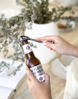 A hand-forged black iron bottle opener in use, effortlessly opening a Burleigh Bighead beer bottle against a stylish neutral-toned setting with a white ceramic vase and dried foliage