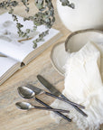 A minimalist table setting featuring hand-forged stainless steel cutlery with sleek black handles, paired with rustic ceramic dishware on a textured stone table, evoking a refined yet organic dining experience.