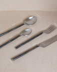 A set of four handcrafted stainless steel cutlery pieces with hand-forged handles, elegantly presented in a minimalist kraft gift box. Includes a dinner knife, fork, tablespoon, and teaspoon for a refined dining experience