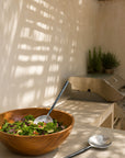 Handcrafted Artisan Salad Servers in forged stainless steel styled with a timber salad bowl in an outdoor kitchen setting with natural light and textured walls
