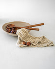 Flatlay of handcrafted wooden salad servers styled in a neutral ceramic bowl with chestnuts and a textured linen cloth on a soft white backdrop.