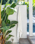 Ivory handloom linen bath towel hanging on an outdoor wall beside tropical foliage and timber decking, capturing a relaxed resort-style atmosphere by the pool.