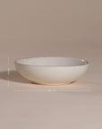 A handmade ceramic bowl with a smooth, speckled neutral glaze, set against a soft beige background. The minimalist design and organic shape make it a versatile piece for modern dining and home decor