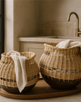 Pair of handwoven rattan baskets with rounded wooden bases, styled with soft neutral linens, placed on a wooden tray in a light-toned laundry room.