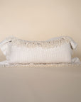 A natural-toned lumbar cushion with a woven texture, featuring delicate fringed edges, set against a neutral backdrop