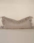 A natural-toned lumbar cushion with a woven texture, featuring delicate fringed edges, set against a neutral backdrop