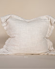 A textured linen cushion in a natural beige tone, featuring a relaxed, oversized design with raw-edge detailing, set against a neutral backdrop