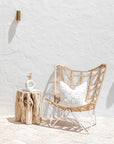 A serene Mediterranean-inspired outdoor space featuring a rattan butterfly chair with a white frame, a natural tree stump side table, and a textured cushion. The open timber doors reveal a breezy indoor lounge with white drapes, a sculptural olive tree in a white planter, and organic wood furniture, bathed in natural sunlight