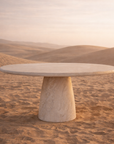 Jabari dining table in dune finish with round tabletop and sculptural tapered pedestal base set in a warm desert landscape.