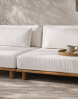 Janelle sofa with low natural timber base and soft white cushions, styled in a warm textured interior with earthy pottery and filtered sunlight.