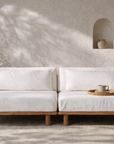 Janelle sofa with low natural timber base and soft white cushions, styled in a warm textured interior with earthy pottery and filtered sunlight.