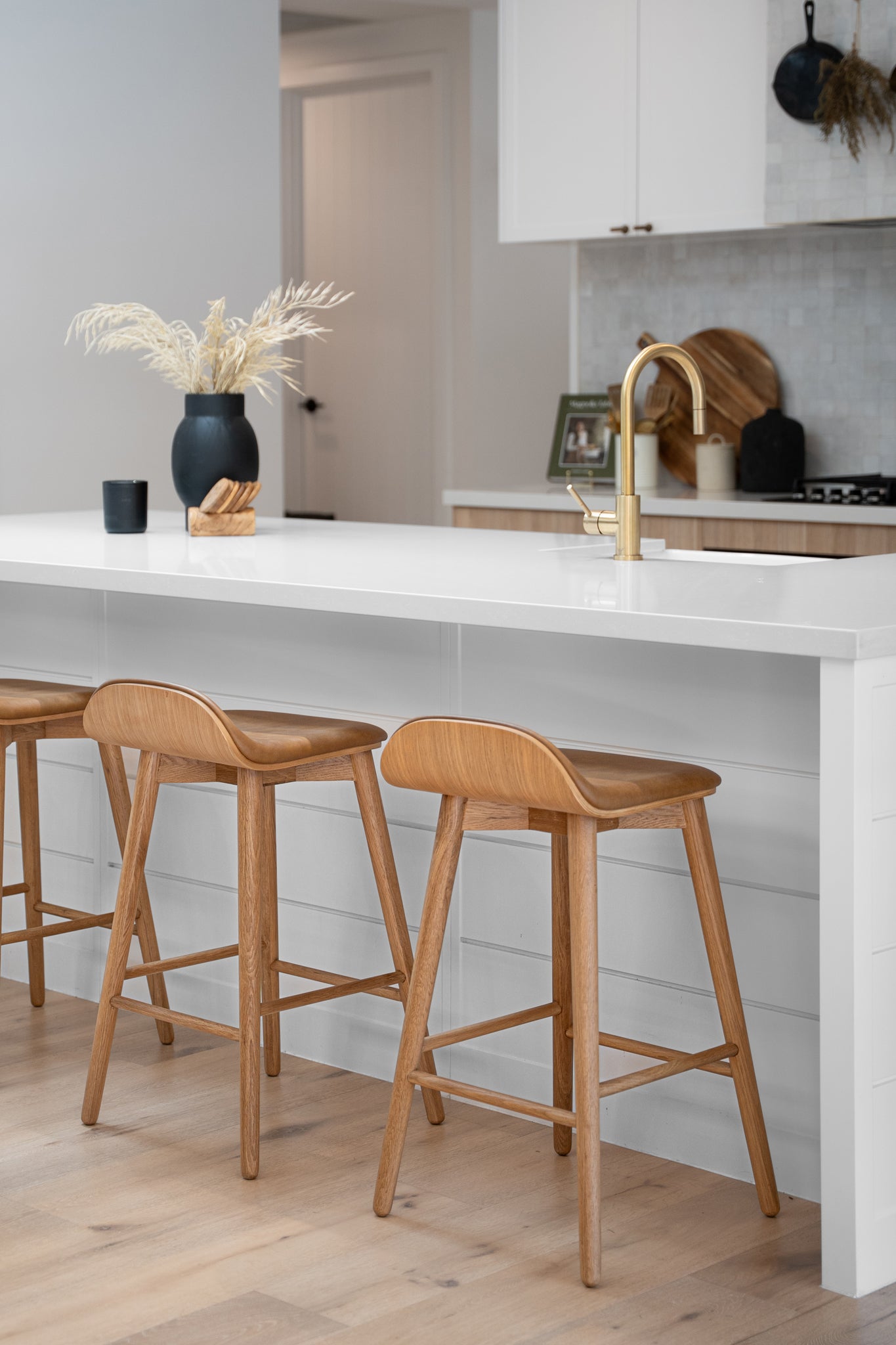 A beautifully styled kitchen with a sleek white island bench, featuring three wooden bar stools with a curved seat design. A modern black vase with dried foliage and a brass faucet add warmth and sophistication to the contemporary space