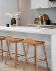 A beautifully styled kitchen with a sleek white island bench, featuring three wooden bar stools with a curved seat design. A modern black vase with dried foliage and a brass faucet add warmth and sophistication to the contemporary space