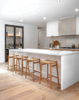 Modern white kitchen featuring KULALA Living teak barstools with woven seats, styled against a crisp island bench and soft timber flooring for a relaxed, coastal feel.