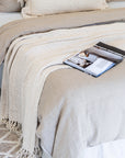 Detail of layered linen bedding and a chunky knit throw in soft neutral tones, styled with an open magazine on a textured wool floor rug