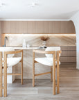 Pair of light oribberwood and boucle counter stools at a marble island bench in a contemporary kitchen with timber cabinetry and natural stone splashback.