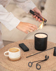 Hand lighting a KULALA Living black soy candle with matchstick, styled on woven table with ceramic coffee cup and candle care tools