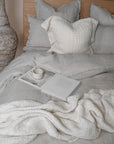 A serene bedroom setting featuring soft grey linen bedding, a handwoven cream throw, and a textured cushion. A ceramic mug rests atop an open book, adding to the relaxed, inviting ambiance