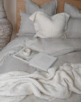 A serene bedroom setting featuring soft grey linen bedding, a handwoven cream throw, and a textured cushion. A ceramic mug rests atop an open book, adding to the relaxed, inviting ambiance