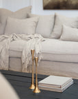 A cosy living space featuring a neutral linen sofa adorned with soft cushions and a textured throw. In the foreground, brass candlesticks and stacked books sit atop a black timber coffee table, creating an elegant yet relaxed styling moment