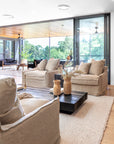 Relaxed living room with natural linen armchairs, black coffee table, and large windows showcasing lush greenery