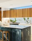 A luxurious modern kitchen featuring a bold black marble island paired with Kulala Living solid oak bar stools. Warm timber cabinetry, white benchtops, and lush greenery create a sophisticated yet inviting space