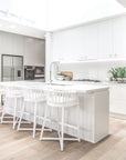 A bright modern kitchen featuring Kulala Living Makena Bar Chairs in crisp white, offering a sculptural rattan backrest and plush seating. Styled with neutral tones, natural wood cabinetry, and fresh greenery for an inviting, coastal-inspired space
