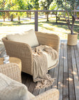 Malusi one seater rattan lounge chair with neutral cushions and textured throw, styled on a timber deck with outdoor side table and olive tree