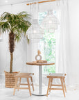 Mara basket styled as a woven planter beside a round timber table with stools in a bright coastal interior with white curtains and rattan pendant lights.