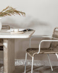 A serene dining setting featuring a marble dining table paired with a woven rope dining chair with a white metal frame. A handmade ceramic espresso cup sits on a matching saucer, complemented by an open book and a white ceramic vase with natural foliage. A perfect blend of organic textures and contemporary elegance