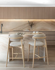 Contemporary kitchen design featuring a marble island with two light ash timber barstools, soft taupe upholstery, and minimal styling in a neutral palette.