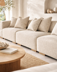 Melonee modular sofa in soft beige upholstery styled in a warm neutral living room with plaster walls, woven rug and round timber coffee table.