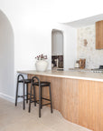 Two KULALA Living Milo Timber Bar Chairs in Espresso styled at a curved timber kitchen island with a natural stone benchtop, textured stone feature wall and warm coastal interiors.