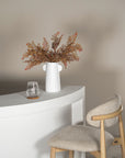 A contemporary wooden barstool with an upholstered seat and curved backrest, positioned at a white curved bar with a minimalist vase and dried foliage arrangement.