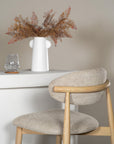 A contemporary wooden barstool with an upholstered seat and curved backrest, positioned at a white curved bar with a minimalist vase and dried foliage arrangement.
