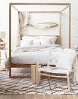 Light-filled modern boho bedroom with a wooden canopy bed, layered neutral bedding, woven accent chair with white rope weave, natural wood side tables, and organic decor accents