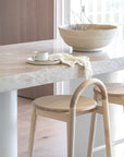 Natural timber stools at a travertine breakfast bar styled with a sculptural ceramic bowl, handmade cup and plate, and soft textured cloth in a light-filled KULALA Living kitchen setting