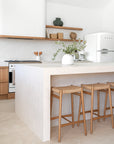 Modern coastal kitchen with a minimalist stone island, woven timber bar stools, open timber shelving, and natural light filtering through skylights.
