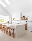 Modern coastal kitchen with a minimalist stone island, woven timber bar stools, open timber shelving, and natural light filtering through skylights.
