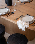 A stylish dining table setting featuring handcrafted ceramic plates, matte black cutlery, and a fringed linen napkin on a natural timber table, complemented by blackwood stools with a textured finish