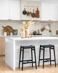 A beautifully styled modern kitchen featuring a white island with black woven bar stools, natural wood cabinetry, and a tiled backsplash. The space is accented with wooden kitchenware, hanging cookware, and sculptural wooden vases, creating a warm and inviting aesthetic with a balance of contemporary and rustic elements