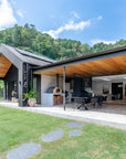 A stylish outdoor dining space featuring a black gable house with timber ceiling details, an open-plan living area, and a built-in wood-fired pizza oven. Surrounded by lush greenery, this modern retreat blends contemporary architecture with natural elements