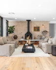 A modern rustic lounge featuring natural linen sofas, a black timber coffee table, and a cozy fireplace set against a textured brick wall. Earthy tones, organic décor, and layered textures create a warm and inviting living space
