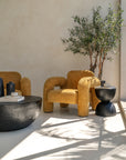 A modern mustard bouclé armchair with curved lines, paired with a round black magnesium coffee table and a sculptural black side table. Styled in a sunlit space with natural shadows and earthy tones, creating a warm and inviting setting