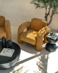 A modern mustard bouclé armchair with curved lines, paired with a round black coffee table and a sculptural black side table. Styled in a sunlit space with natural shadows and earthy tones, creating a warm and inviting setting.