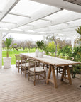 An inviting outdoor dining setting featuring natural rattan dining chairs with plush cushions, paired with a rustic timber table under a white pergola, overlooking a scenic orchard landscape.