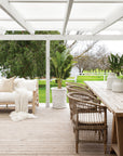 An inviting outdoor dining setting featuring natural rattan dining chairs with plush cushions, paired with a rustic timber table under a white pergola, overlooking a scenic orchard landscape